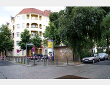 Straßenecke mit einem hellgelben Wohngebäude im Altbaustil, verziert mit halbrunden Balkonen und einem roten Ziegeldach; umgeben von Bäumen, einer Litfaßsäule und parkenden Autos. Ein Straßenschild markiert den „Böhmischen Platz“, während Verkehrsschilder und Absperrungen auf die Verkehrsführung hinweisen.