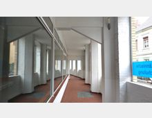 Ein überdachter, heller Gang mit weißen Wänden, rechteckigen Fenstern und roten Fliesenboden. Links befindet sich eine Glasfassade mit Spiegelungen, rechts sind weiße Backsteinwände und ein teilweise sichtbares blaues Schild.