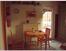 Kleine Küchen-Ecke mit Holztisch und zwei Holzstühlen, einem karierten Tischläufer und einer Vase mit Blumen. Im Hintergrund eine Wand mit Heizung, Wanddekorationen, einem Regal und eine Glastür mit Blick nach draußen.