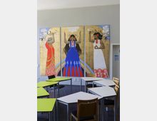 Ein Klassenraum mit modernen Schulmöbeln, darunter Tische mit grünen und weißen Tischplatten sowie Stühle. An der Wand hängt ein dreiteiliges Wandgemälde mit stilisierten Darstellungen von drei Frauen in traditionellen Kleidern vor einem goldenen Hintergrund, umgeben von symbolhaften Elementen wie Blitzen, Blumen und Regenbögen.