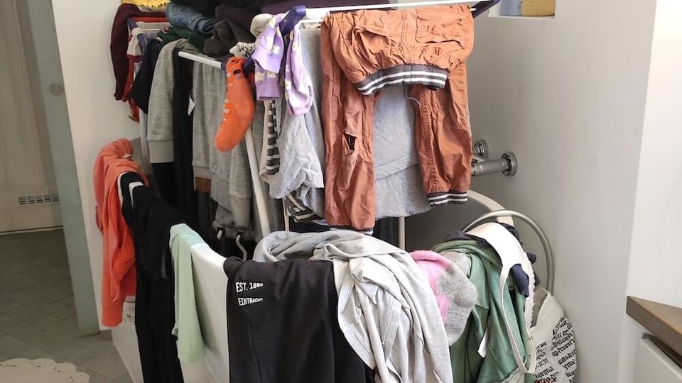 A bathroom with a white bathtub being used as a drying area for clothes, featuring a portable drying rack filled with garments. Above the tub is a wall-mounted water heater, and recessed shelves contain personal care products.