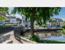 Kleiner Park mit einem Baum im Vordergrund, einer Fußgängerbrücke über einen Fluss und einer Reihe von Wohnhäusern im Hintergrund. Die Umgebung umfasst gepflasterte Wege, Wasser sowie Parkplätze mit Fahrrädern und Autos.