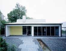 Ein modernes, einstöckiges Gebäude mit Flachdach und großen Fenstern auf der rechten Seite. Der Vorhof ist mit grauem Kies und rechteckigen Steinplatten gestaltet, ergänzt durch einen schmalen, flachen Wassergraben. Im Hintergrund sind Bäume sichtbar.