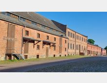 Ziegelstein-Gebäudekomplex mit historischen Lager- und Verwaltungsbauten, rechteckigen Fenstern und Holztüren, flankiert von Kopfsteinpflaster und Grünflächen, unter einem klaren blauen Himmel.