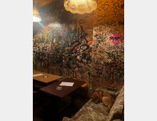 Innenraum eines Cafés oder einer Bar mit zwei kleinen Holztischen und gedämpfter Beleuchtung durch hängende Lampenschirme. Die Wände sind vollständig mit dicht übereinanderliegenden Graffiti-Tags, Kritzeleien und Stickern bedeckt, daneben steht ein gemustertes Sofa.