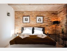 Schlafzimmer mit einem Doppelbett, umgeben von einer Backsteinwand. Auf dem Bett liegen braune Decken und gefaltete Handtücher; seitlich stehen Nachttische mit kleinen Lampen, ein Stehlampe und ein Schreibtisch, minimalistisch dekoriert.