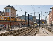 Straßenszene mit einer erhöhten roten Backsteinbrücke, auf der eine gelbe S-Bahn fährt, und darunter Fußwege und Ladenfronten. Im Vordergrund verlaufen Straßenbahnschienen mit einer gelben Straßenbahn, flankiert von Gehsteigen, Fahrrädern und städtischer Architektur.