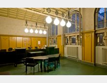 Innenraum eines historischen Büros im Jugendstil mit hohen Decken, verzierten Holzwänden und großen Fenstern, die Buntglasdetails aufweisen. Im Raum befinden sich moderne Büromöbel, ein Kronleuchter mit kugelförmigen Lampen, sowie Heizkörper unter den Fenstern.
