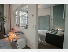 Modernes Apartment mit einem offenen Raumkonzept, das ein Schlafzimmer mit einem Bett und gepolstertem Kopfteil sowie einen Wohnbereich mit Metallregalen und einem Plattenspieler zeigt. Eine Glaswand mit mehreren kleinen Fenstern trennt die Räume und lässt viel Licht herein.