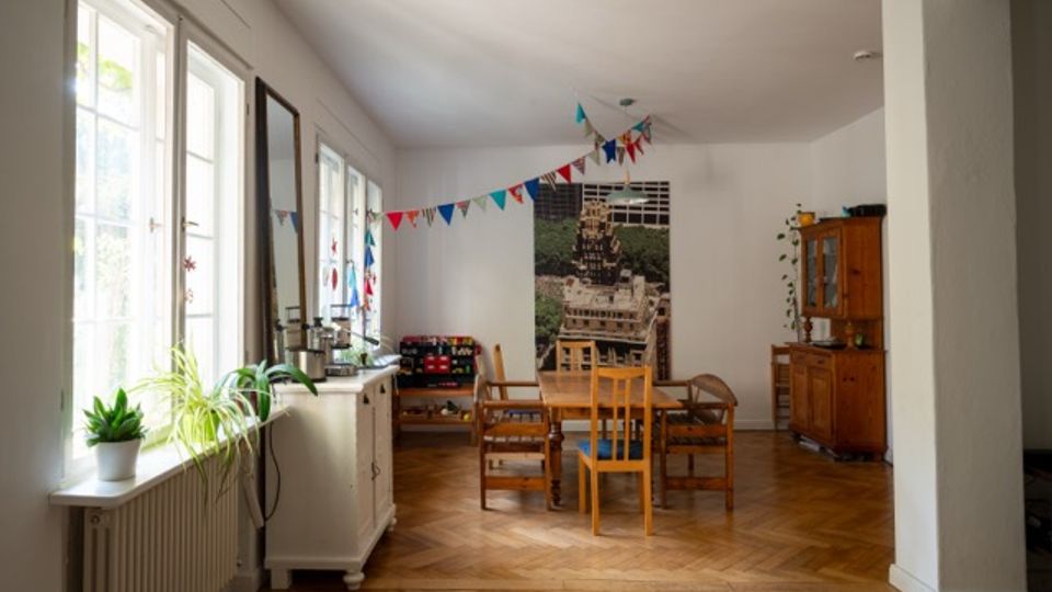 Heller Wohn- und Essbereich mit großen Fenstern entlang der linken Wand, Holzboden im Fischgrätmuster und einem Tisch mit Stühlen in der Raummitte. An der Rückwand hängt ein großes Wandbild, darüber eine bunte Wimpelgirlande; rechts steht ein Holzsideboard mit Pflanzen.