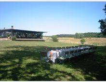 Ein moderner Pavillon mit großen Glasflächen steht auf einer weitläufigen Wiese, umgeben von Weiden und Bäumen. Im Vordergrund ist eine lange, festlich gedeckte Tafel mit Stühlen unter freiem Himmel zu sehen.