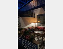 Gemütlicher Sitzbereich in einem Wintergarten bei Nacht, mit einem Sofa und Kissen, einem Glastisch, dekorativen Pflanzen sowie einer Stehlampe, die warmes Licht verbreitet. Der Raum hat schwarz-weiß karierte Fliesen und ein Oberlicht mit floralem Muster.