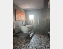 Modernes Badezimmer mit freistehender weißer Badewanne auf einer erhöhten Plattform aus grauen Fliesen. Der Boden ist mit gemusterten schwarz-weißen Fliesen ausgelegt, ein Fenster sorgt für natürliches Licht, und im Raum befinden sich Dekorationen wie ein bunter Kunstdruck und eine Pflanze.