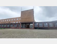 Backsteinbau mit flachem Dach und großem zentralen, turmartigem Abschnitt, umliegenden rechteckigen Fenstern und einer Eingangsfront mit Glasbaustein-Elementen; die Fassade öffnet sich zu einem gepflasterten Vorplatz unter bewölktem Himmel.