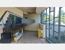 Innenansicht eines modernen Gebäudes mit großer Fensterfront, die nach außen auf eine grüne Umgebung blickt. Im Vordergrund eine schwarze, geschwungene Treppe mit Geländer; der Raum weist sichtbare Gebrauchsspuren und abblätternde Farbe an der Decke auf.