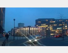 Abendaufnahme des Bahnhofs Potsdamer Platz in Berlin, umgeben von modern beleuchteten Bürogebäuden. Im Vordergrund ist eine Straße mit fahrendem Verkehr zu sehen, während hinter dem Bahnhof die Fenster der Gebäude hell erleuchtet sind.