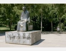 Bronzestatue einer sitzenden Person auf einem rechteckigen Sockel, umgeben von Bäumen in einem offenen Parkbereich. Der Sockel ist mit Graffiti bedeckt, und im Hintergrund sind große Nadelbäume und ein Mülleimer sichtbar.