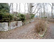 Gepflegte Garten- oder Parkanlage mit einem geschwungenen Weg, der von niedrigen Steinmauern und Büschen gesäumt ist. Im Hintergrund sind kahle Bäume und eine leicht sichtbare Bahnstrecke erkennbar.