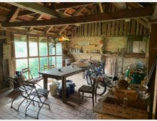 Rustikaler Innenraum einer Scheune oder Werkstatt mit Holzbalken und Ziegeldach. Im Vordergrund ein Holztisch mit Stühlen, im Hintergrund Fahrräder, Werkzeuge, dekorative Gegenstände und ein Fenster mit Blick auf grüne Vegetation.