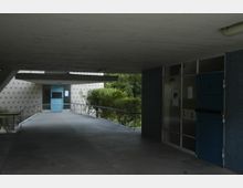 Überdachter Zugang zu einem Gebäude mit grauem Betonboden und rechteckigen, blauen Türen. Die Fassade hat ein Punktmuster, daneben befinden sich eine Rampe mit Geländer und Büsche im Hintergrund.