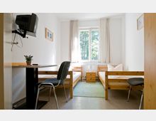 Ein kleines, schlicht eingerichtetes Zimmer mit zwei Einzelbetten aus Holz, einem kleinen Tisch mit Stuhl und einem an der Wand montierten Fernseher. Ein Fenster mit hellen Vorhängen lässt Tageslicht herein, und unter dem Fenster befindet sich ein Heizkörper.