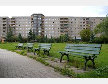 Ein Wohnblock mit mehreren Etagen und Balkonen im Hintergrund, davor eine Grünfläche mit Bäumen und Blumen. Im Vordergrund stehen mehrere grüne Parkbänke neben einem gepflasterten Weg.