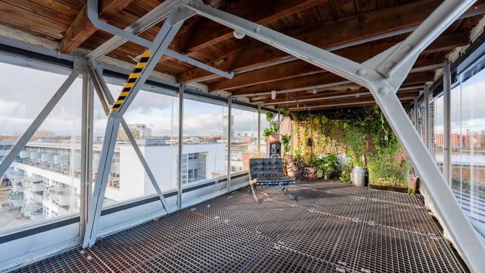 Überdachte Dachterrasse mit Stahlträgerkonstruktion, Holzbalkendecke und Gitterrostboden, umgeben von großen Glasfronten mit Blick auf umliegende Gebäude. In einer Ecke befindet sich eine begrünte Pflanzenwand mit Topfpflanzen.