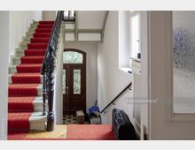 Treppenhaus eines Altbaus mit Holztreppe, die mit rotem Teppich belegt ist, und einem dekorativen schwarzen Geländer. Im Hintergrund eine Holzeingangstür mit Glaseinsätzen und Metallgittern, daneben ein kleines Fenster und Gegenstände im Eingangsbereich.