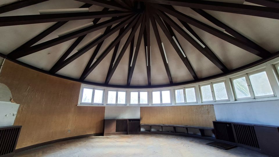 Innenraum eines runden Pavillons mit kuppelförmiger Decke, deren dunkle Balken strahlenförmig zur Mitte verlaufen. Entlang der gebogenen Wand befindet sich ein umlaufendes Band aus kleinen Fenstern über einer niedrigen Sitzbank und Lüftungsgittern.