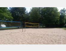 Sandplatz mit mehreren Volleyballnetzen in einem Park, umgeben von grünen Bäumen. Im Hintergrund ist ein weißes Zelt zu sehen.