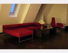 Modern eingerichteter Wohnraum mit schräger Dachwand und einem Dachfenster. Ein rotes Sofa und ein passender Sessel stehen auf dunklem Holzboden, daneben ein niedriger Couchtisch mit einer Vase mit Blumen und einer Wasserflasche.