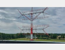 Großer rot-weißer Sendemast mit mehreren horizontalen und schrägen Streben, umgeben von Grünflächen und Bäumen, unter einem bewölkten Himmel.