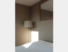 Minimalistisches Schlafzimmer mit neutralen Wandfarben, einem weißen Bett, einer Stehlampe mit hellem Lampenschirm und einem rechteckigen Wandspiegel mit Holzrahmen. Sonnenlicht fällt schräg durch ein Fenster auf die Wand und das Bett.