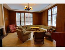 Gemütlicher Wohnraum mit Holzwänden, großen Fenstern und Blick auf einen verschneiten Wald. Eine Sitzgruppe mit verschiedenen Sesseln, einem geblümten Ecksofa und einem runden Tisch steht auf einem gefliesten Boden; links ist ein Kamin sichtbar.