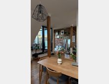 Wohnraum in einem modernen Loft mit sichtbaren Backsteinwänden, Holzbalken und großen Fenstern. Im Vordergrund befindet sich ein Holzesstisch mit einem Blumenstrauß in einer weißen Vase, im Hintergrund eine offene Sitzecke mit dekorativen Regalen.