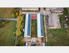 Luftaufnahme eines landwirtschaftlichen Hofs mit zwei langen Gebäuden, deren Dächer vollständig mit Solarpaneelen bedeckt sind. Zwischen den Gebäuden befindet sich ein bewachsener Innenbereich, umgeben von Grünflächen, Bäumen und weiteren Gebäuden mit teils eingelagerten Materialien.