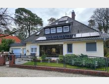 Zweistöckiges Einfamilienhaus mit dunkelgrauem Satteldach, auf dem Solarpaneele installiert sind. Das Haus hat eine helle Fassade, große Fenster, einen kleinen Garten mit Zaun sowie eine gepflasterte Einfahrt neben einer Garage.