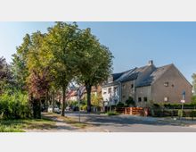 Straßenansicht einer ruhigen Wohngegend mit Einfamilienhäusern in Reihenanordnung, umgeben von Bäumen und gepflegten Gärten. Am rechten Bildrand ist ein Gehweg mit Straßenschild und ein Parkplatz für Menschen mit Behinderung zu sehen.