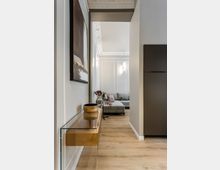 Blick durch einen schmalen Flur mit hellem Holzboden und weißer Wandvertäfelung in ein Wohnzimmer mit grauem Ecksofa. An der linken Wand hängt ein großes abstraktes Bild über einer schwebenden Glas-Konsole, rechts steht ein hoher dunkler Schrank; an der Decke sind Stuckleisten und eine Wandleuchte zu sehen.