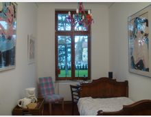 Kleines, schlichtes Schlafzimmer mit einem Holzbett, einem gepolsterten Stuhl in Karomuster und einem großen Fenster mit Blick auf eine grüne Rasenfläche und Bäume. An den weißen Wänden hängen moderne, farbenfrohe Gemälde, und ein farbiger Kronleuchter hängt von der Decke.