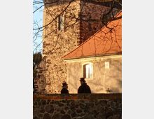 Historische Kirche aus Naturstein mit rotem Ziegeldach, im Vordergrund zwei Personen in traditioneller Kleidung hinter einer Mauer, umrahmt von kahlen Ästen.