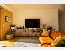 Wohnzimmer mit Holzboden und heller Wand, ausgestattet mit einem großen Flachbildfernseher, der auf einem Regal steht, das mit Schallplatten gefüllt ist. Im Vordergrund befinden sich ein orangefarbenes Ledersofa mit gelben Kissen und ein heller Teppich.