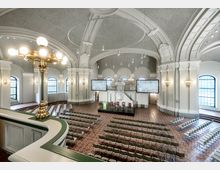 Ansicht eines historischen Saals mit hohen Gewölbedecken, verzierten Säulen und großen Rundbogenfenstern. Der Raum ist mit Stuhlreihen, einem Podium für Redner und Hängelampen ausgestattet, im Hintergrund befindet sich eine geschmückte Kanzel, flankiert von zwei großen Bildschirmen.