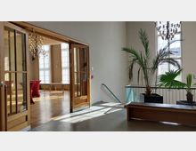 Innenansicht eines lichtdurchfluteten Flurs mit großen Fenstern, einer Holz-Sitzbank, Topfpflanzen und einem Geländer aus Metall. Im Hintergrund ist ein angrenzender Raum mit Parkettboden, Kronleuchtern und einem offenen Holztürrahmen zu sehen.