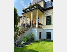 Gelbes Wohnhaus mit historisch anmutenden Säulen und einer Treppe, die zu einer Terrasse mit Blumenkästen führt. Vor dem Haus befindet sich ein gepflegter Garten mit Rasen und Blumenbeeten.