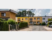 Reihenhäuser in einer Wohnsiedlung mit gelben und weißen Fassaden, Vorgärten mit Hecken und Gehweg. Im Hintergrund sind Bäume und bewölkter Himmel zu sehen.