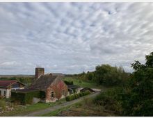 Ländliche Szene mit einem kleinen, verwitterten Steingebäude, bewachsen mit Efeu, daneben ein alter gemauerter Turm. Im Hintergrund sind Felder, Bäume und ein bewölkter Himmel zu sehen.