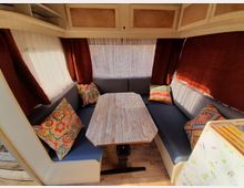 Eine Sitzecke in einem Wohnwagen mit L-förmigen Polsterbänken, einem Holztisch und bunten gemusterten Kissen. Die Fenster sind mit weißen Gardinen und rotbraunen Vorhängen versehen, das Interieur hat helle Holz- und Stoffelemente.