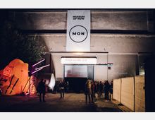 Außenansicht eines Gebäudes mit einem großen Banner "Museum of Now (MON)" über dem Eingang, flankiert von Kunstinstallationen wie einer gelben, beleuchteten Skulptur. Mehrere Personen stehen vor dem Eingang, das Schild weist auf die "Berlin Edition 2019" hin.
