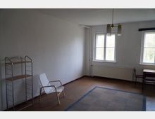 Ein schlicht eingerichteter Raum mit weißen Wänden und einem braunen Bodenbelag. Der Raum enthält ein Regal, einen weißen Stuhl, einen kleinen Tisch mit Stuhl und eine Hängelampe an der Decke; zwei Fenster lassen Tageslicht herein.